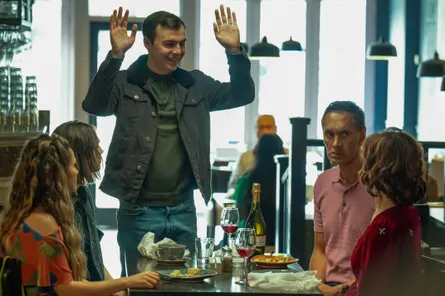 A man stands with his hands raised around a dining table, where four people are seated